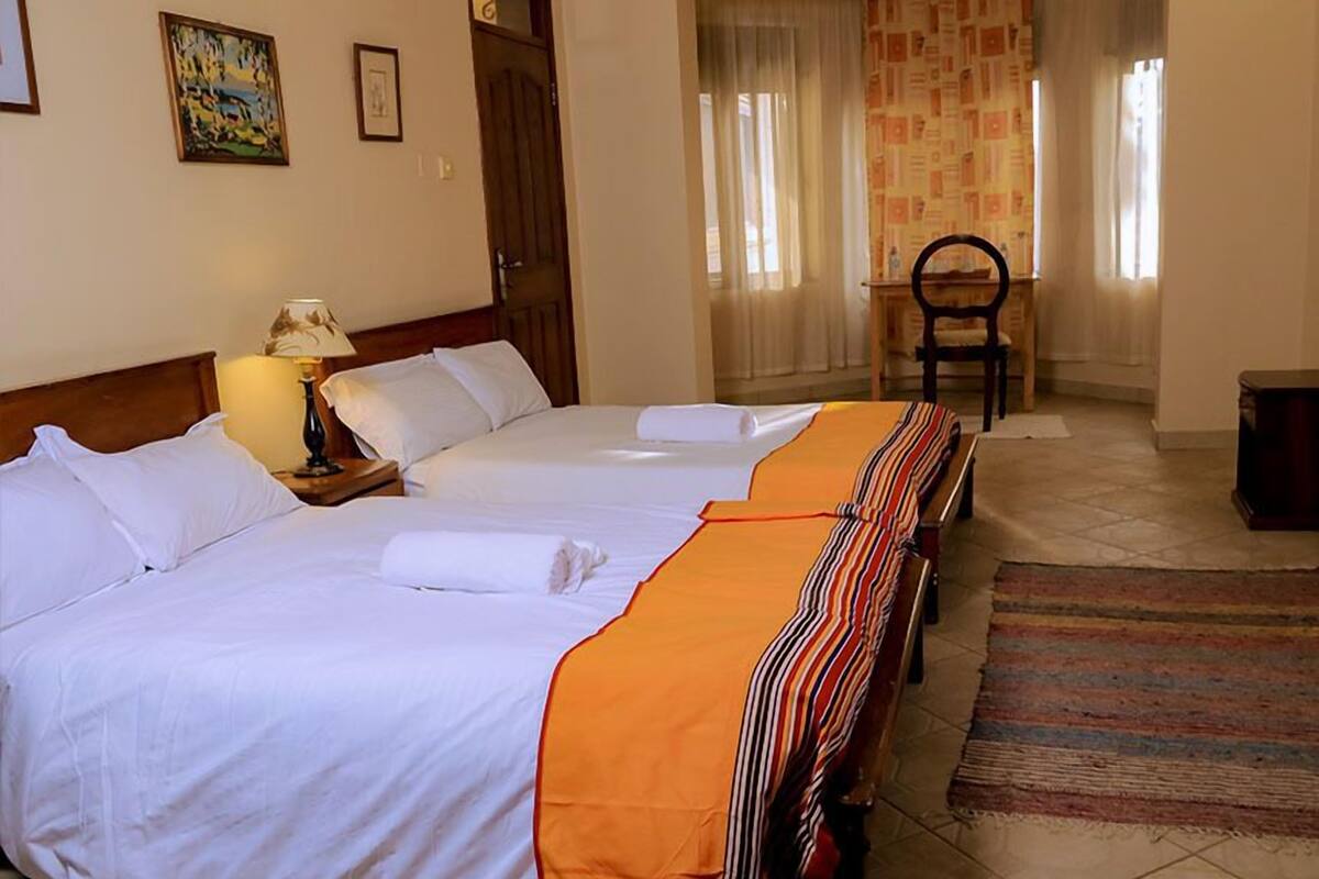 A hotel room with two beds, a wooden headboard, a nightstand with a lamp, a chair, and a view of the hallway.