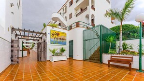 Un patio con suelo de baldosas, un banco, plantas en macetas, una reja y un edificio con un letrero.