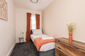 Ein ordentlich eingerichtetes Schlafzimmer mit einem Bett, einem Nachttisch, einer Lampe, einer Kommode und einer Vase mit Blumen.