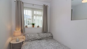 Um quarto pequeno com cama, mesa de cabeceira, abajur, janela com cortinas e uma planta em vaso.