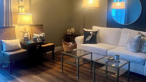 Una sala de estar con un sofá blanco, una mesa de centro de vidrio, sillas de mimbre, una lámpara, un espejo y piso de madera.