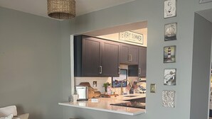 Una cocina moderna con armarios oscuros, encimera blanca y una lámpara colgante. Hay dos taburetes y una mesa de centro en la zona de estar.