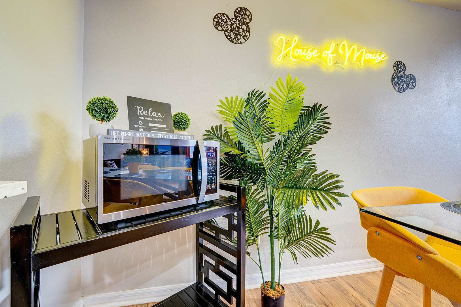 Une cuisine moderne équipée d’un four à micro-ondes, d’une petite plante en pot et d’un panneau lumineux au néon sur lequel on peut lire : « House of Mouse ».