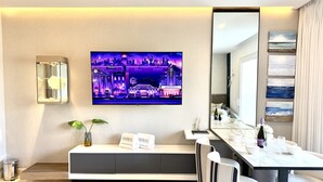 Een moderne hotelkamer met een grote flatscreen televisie aan de muur, een zithoek met een bank en een klein tafeltje, en een eethoek met een lange tafel en stoelen.