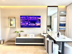 Une chambre d’hôtel moderne dotée d’un téléviseur à écran plat fixé au mur, d’un coin salon avec un canapé et une petite table, ainsi que d’un coin repas avec une longue table et des chaises.