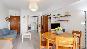 A modern living room with a sofa, a dining table with chairs, a coffee table, a wooden floor, a ceiling light, and a kitchen visible through an open door.