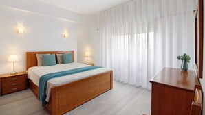 A bedroom with a wooden bed, bedside lamps, a nightstand, a desk, and a window with sheer curtains.