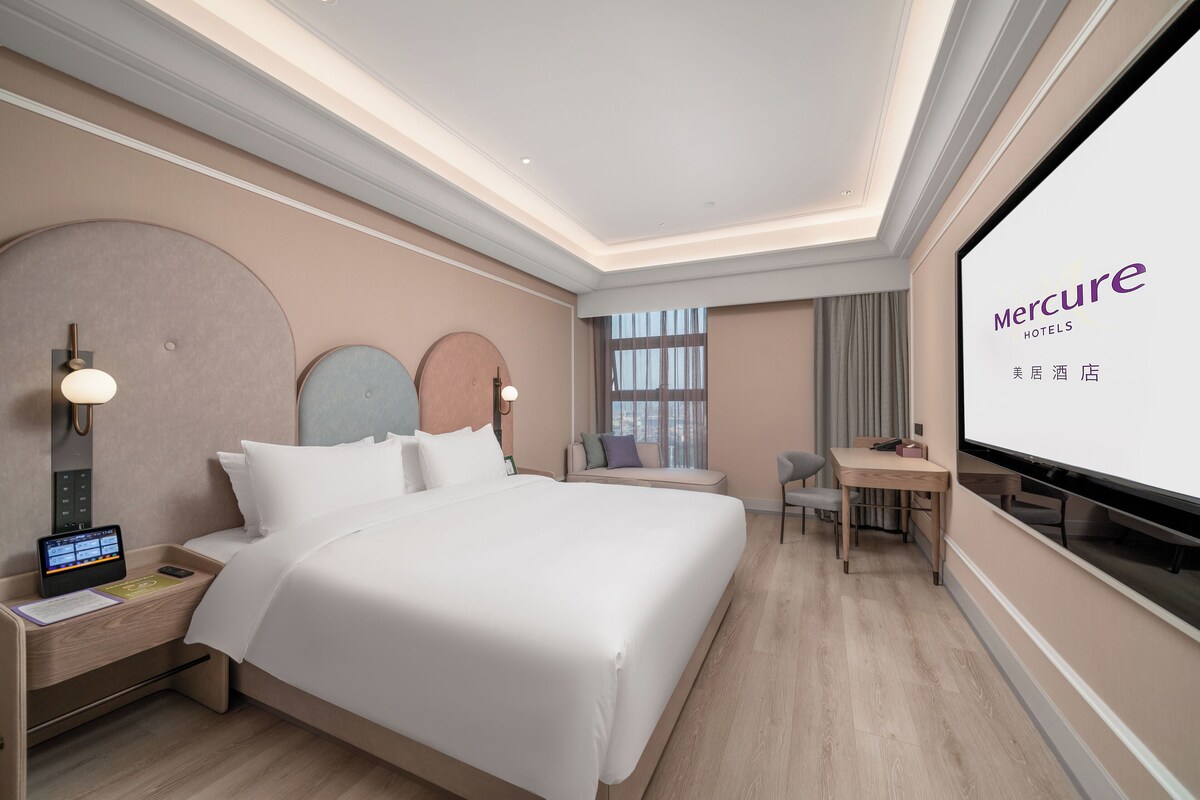 A hotel room with a bed, bedside table, desk, chair, and a large screen displaying the hotel name.