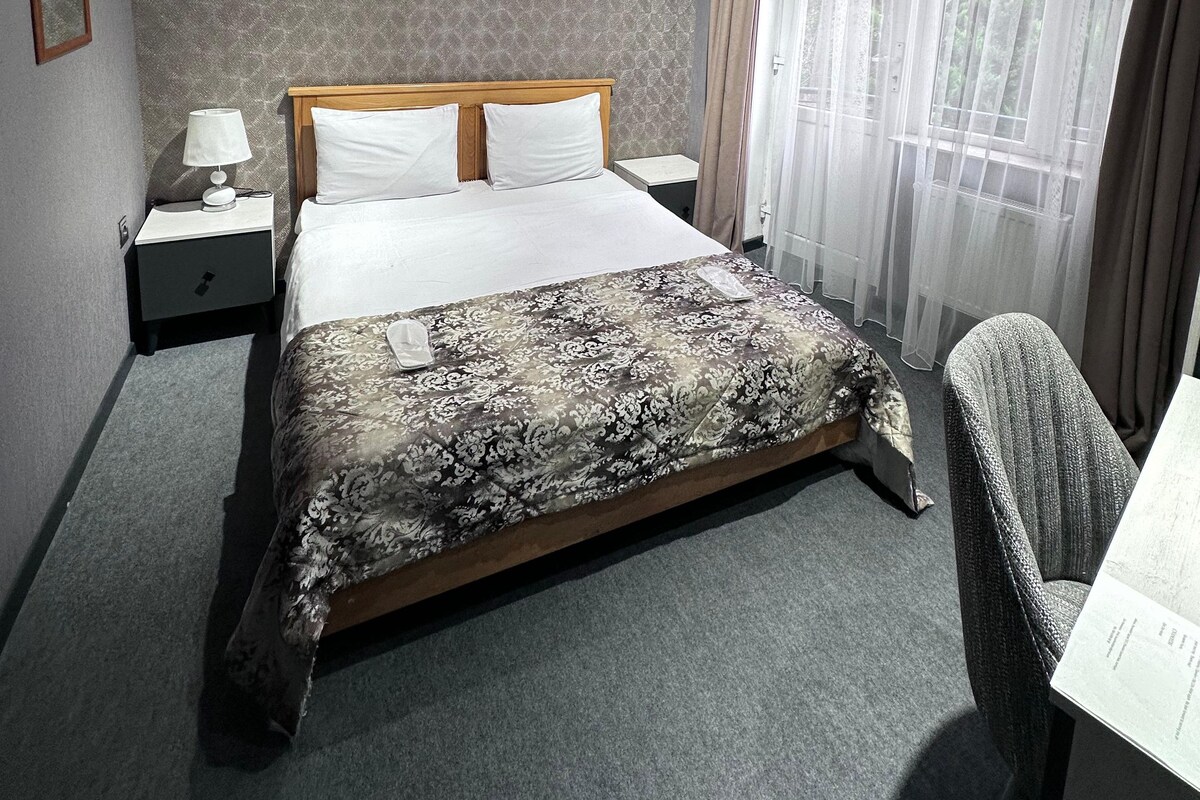 Une chambre d’hôtel comprenant un lit, des tables de chevet, une chaise, un tableau, un climatiseur et une fenêtre avec des rideaux.