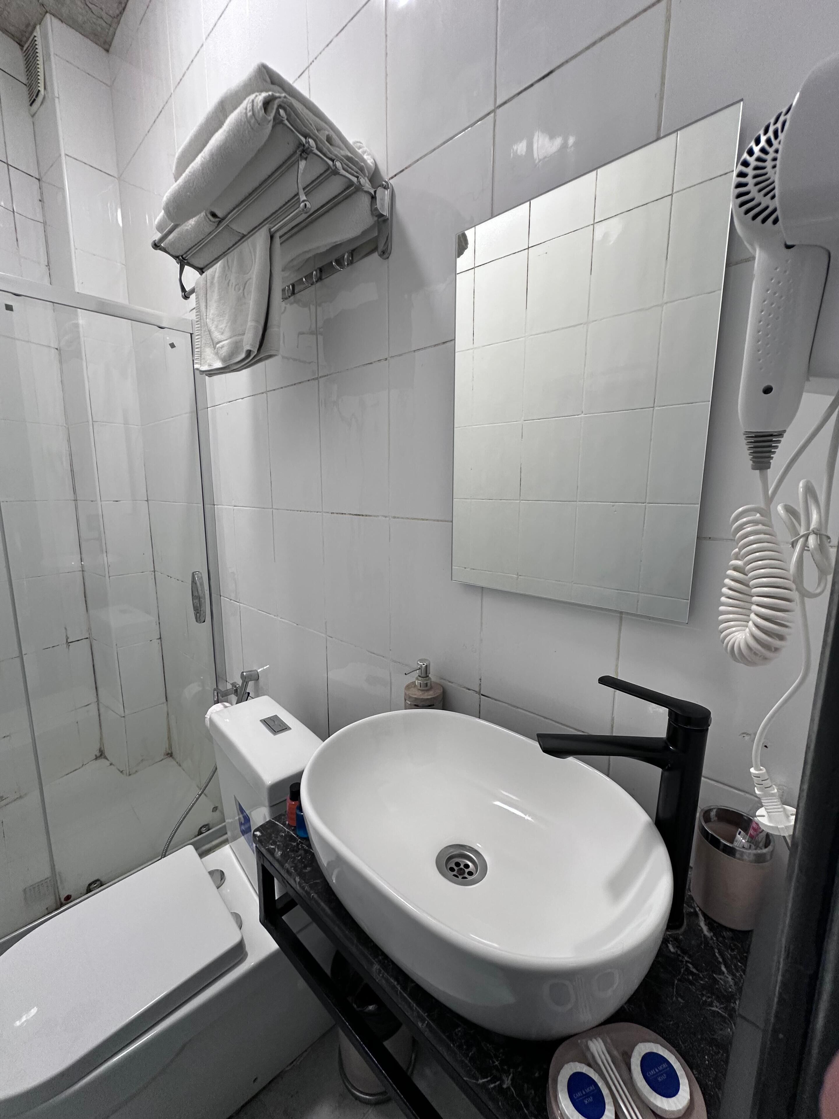 Een badkamer met een wit wastafel, toilet en douche. Boven het wastafel bevindt zich een handdoekhouder en aan de muur hangt een spiegel.