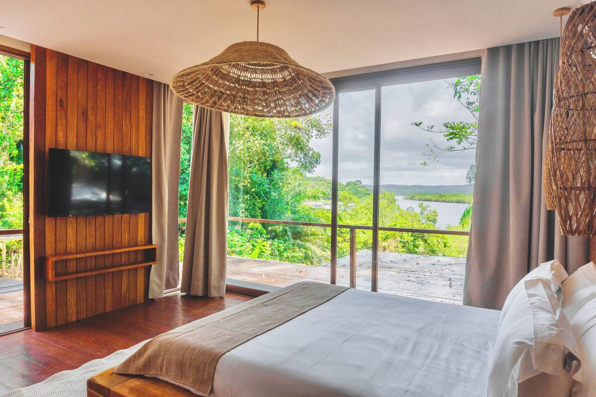A bedroom with a large bed, a flat-screen TV mounted on a wooden panel wall, and a large window offering a view of greenery and a body of water.