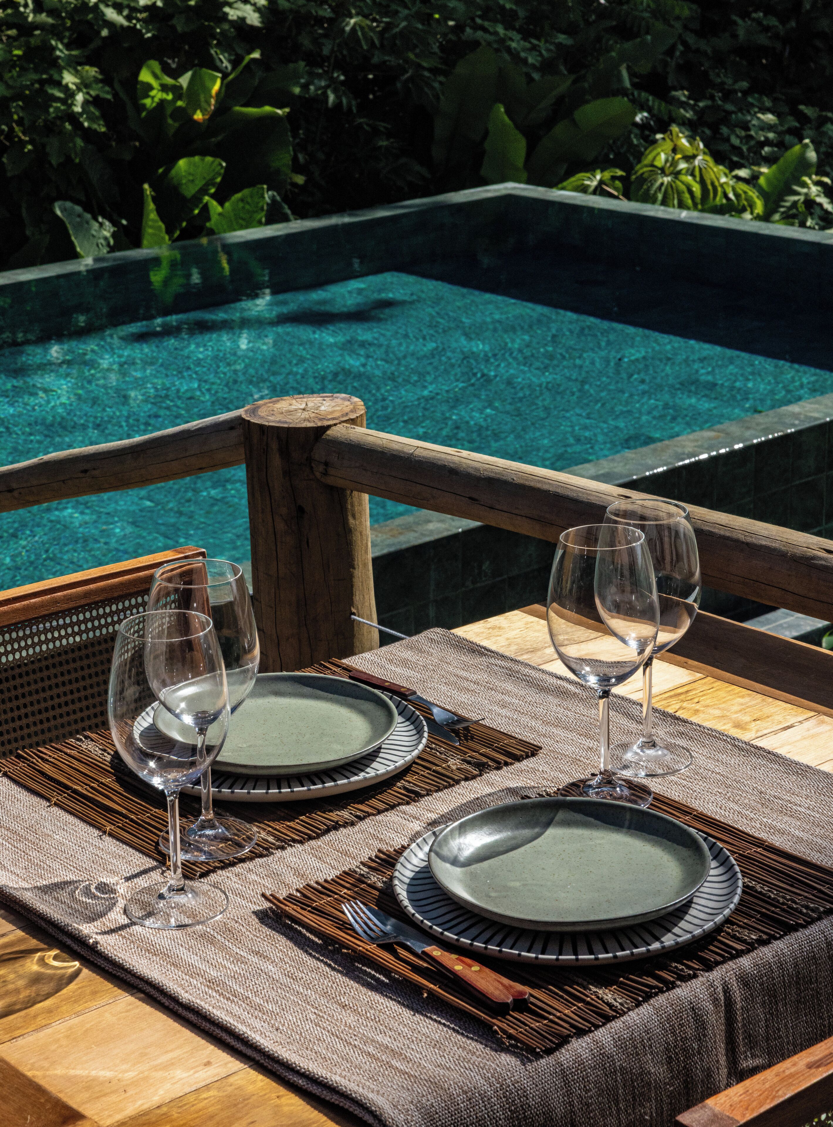 A dining table with plates and wine glasses, overlooking a clear pool.