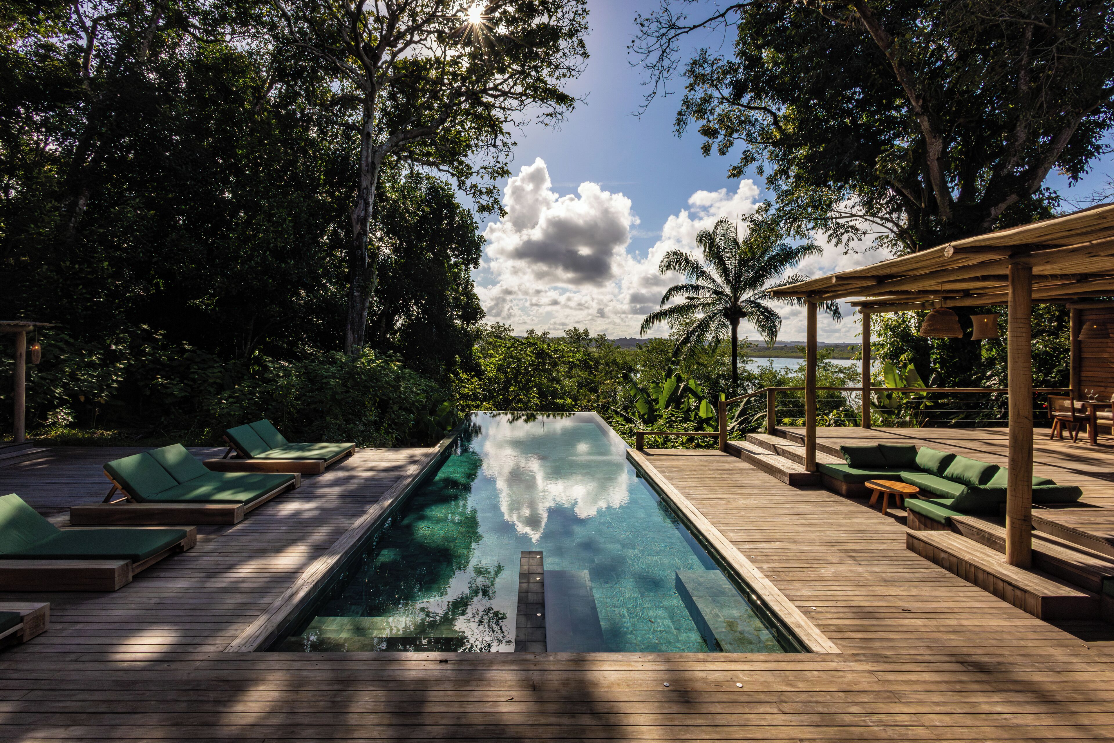 A wooden deck with a pool, green lounge chairs, and a covered seating area with a view of the surrounding greenery and sky.