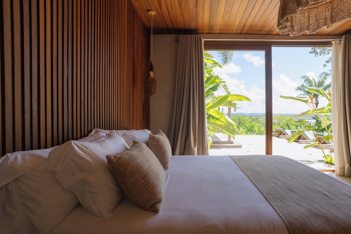 Habitació d'hotel amb llit, cortines, una làmpada penjant i vista a un paisatge tropical.