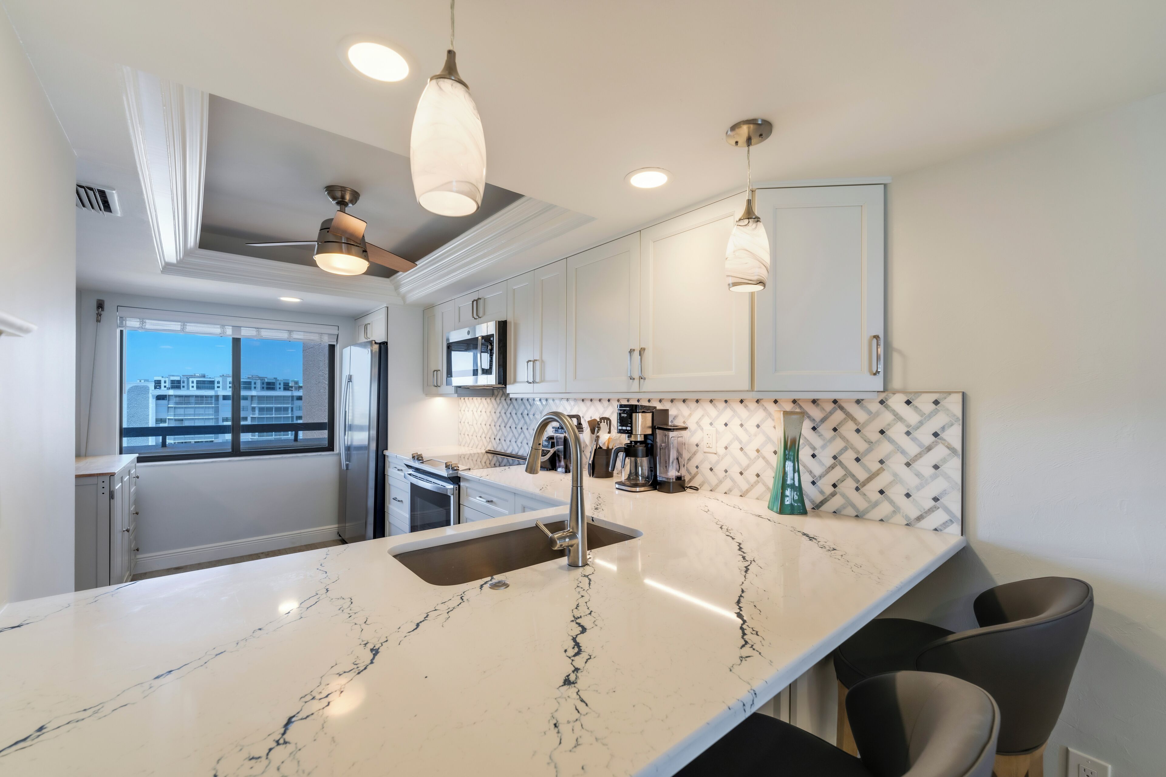 A modern kitchen with a marble countertop, stainless steel appliances, pendant lighting, and a view of the city through the window.