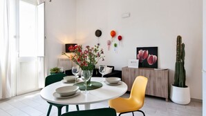 A dining area with a round table, green chairs, and a yellow chair. There is a vase with flowers, a clock, and a cactus on the wall.