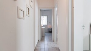 A narrow hallway with white walls and a tiled floor, leading to a room with a window and a small table with a lamp.