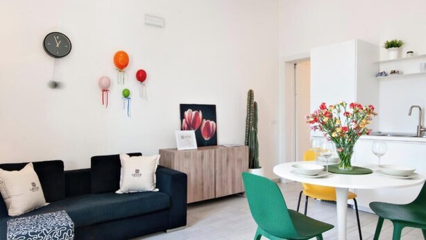 A modern living room with a dark blue sofa, a small table with green chairs, and a white dining area. There is a clock on the wall and a cactus on the windowsill.