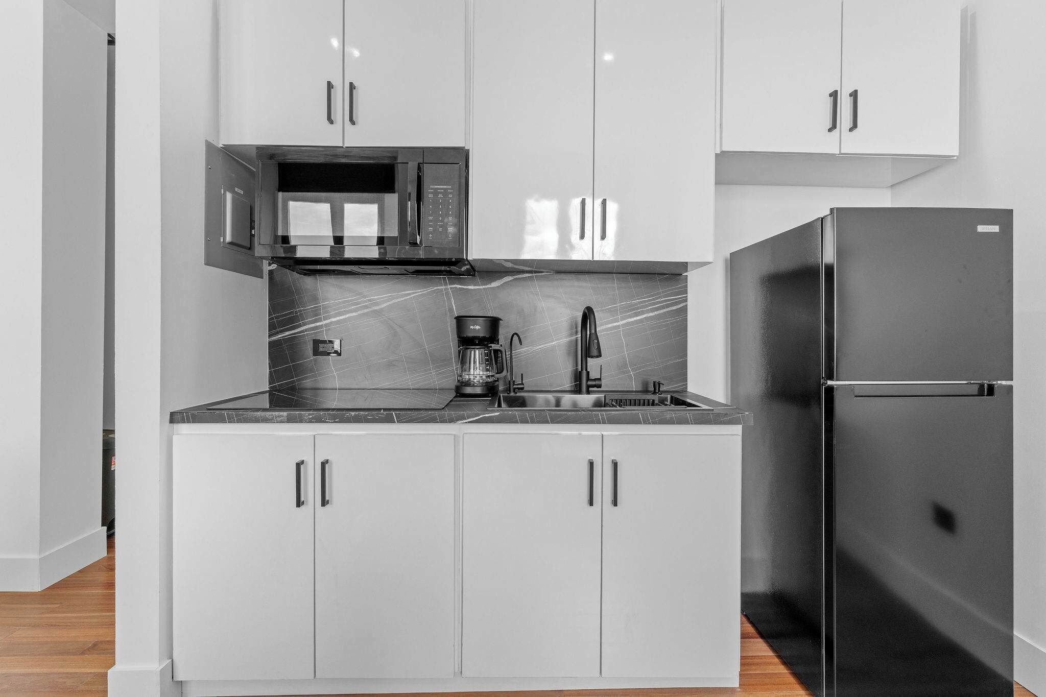 Una cocina compacta con armarios blancos, un refrigerador negro, un microondas y una cafetera.