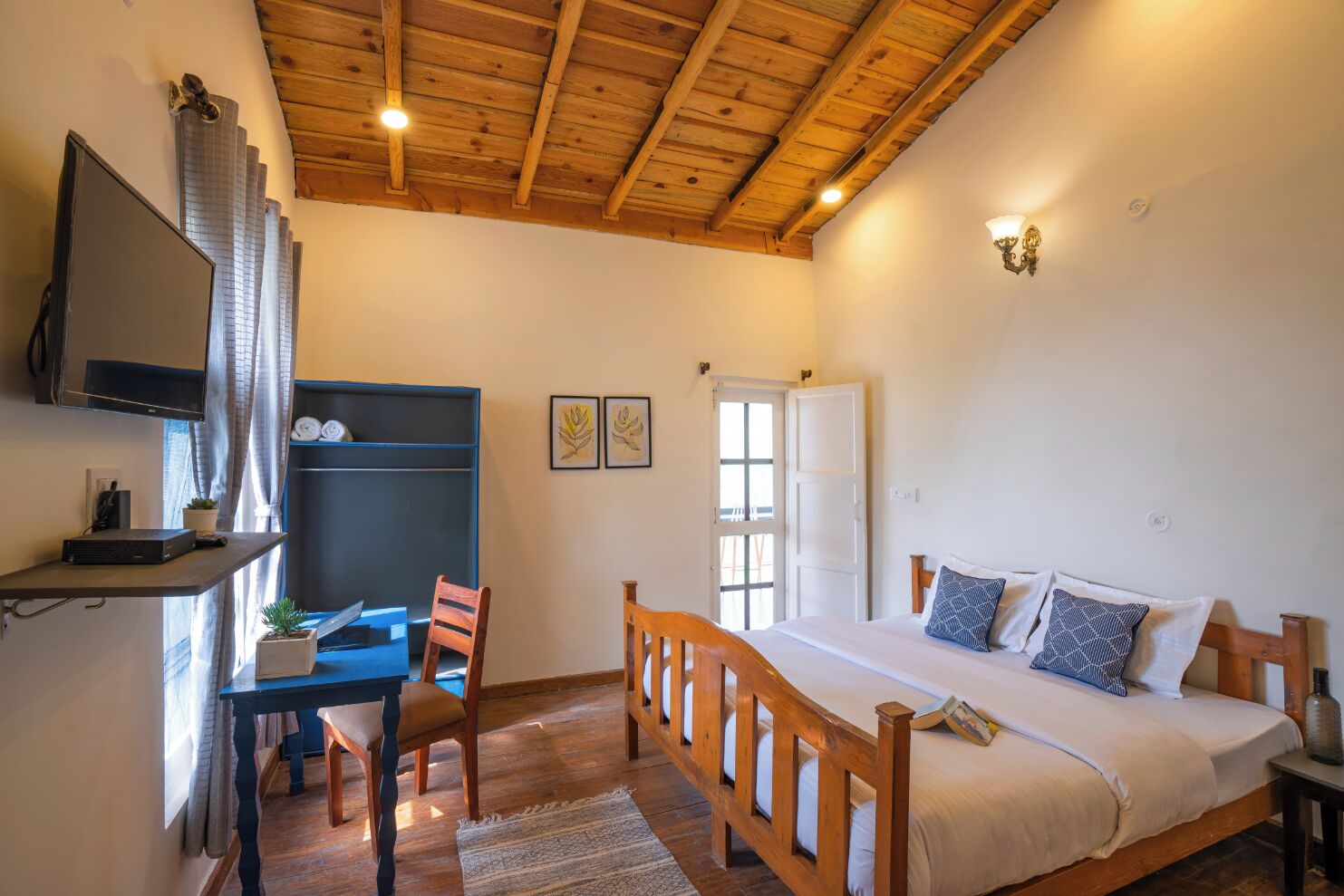 A bedroom with a wooden ceiling, a bed with pillows, a desk with a computer, and a TV mounted on the wall.