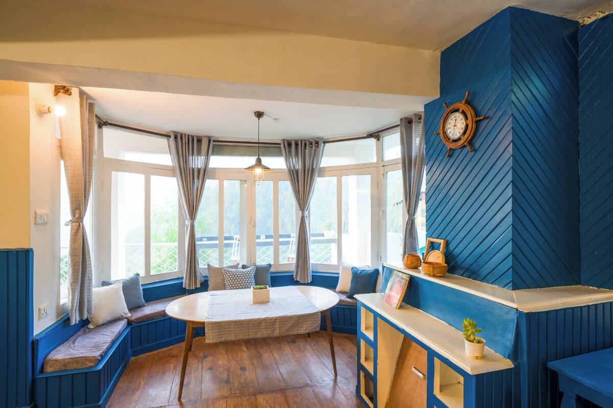 Une pièce avec une table ronde, des bancs bleus, un plafond en bois et un gouvernail de bateau accroché au mur.