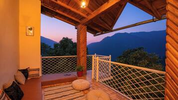 Un balcón de madera con vistas a montañas al atardecer, que incluye una zona de estar con cojines y una pequeña mesa.