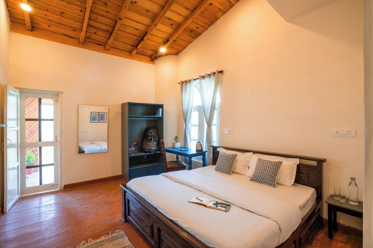 Une chambre avec un plafond en bois, un lit agrémenté d’oreillers à motifs, un bureau avec une chaise, une armoire et une fenêtre avec des rideaux.