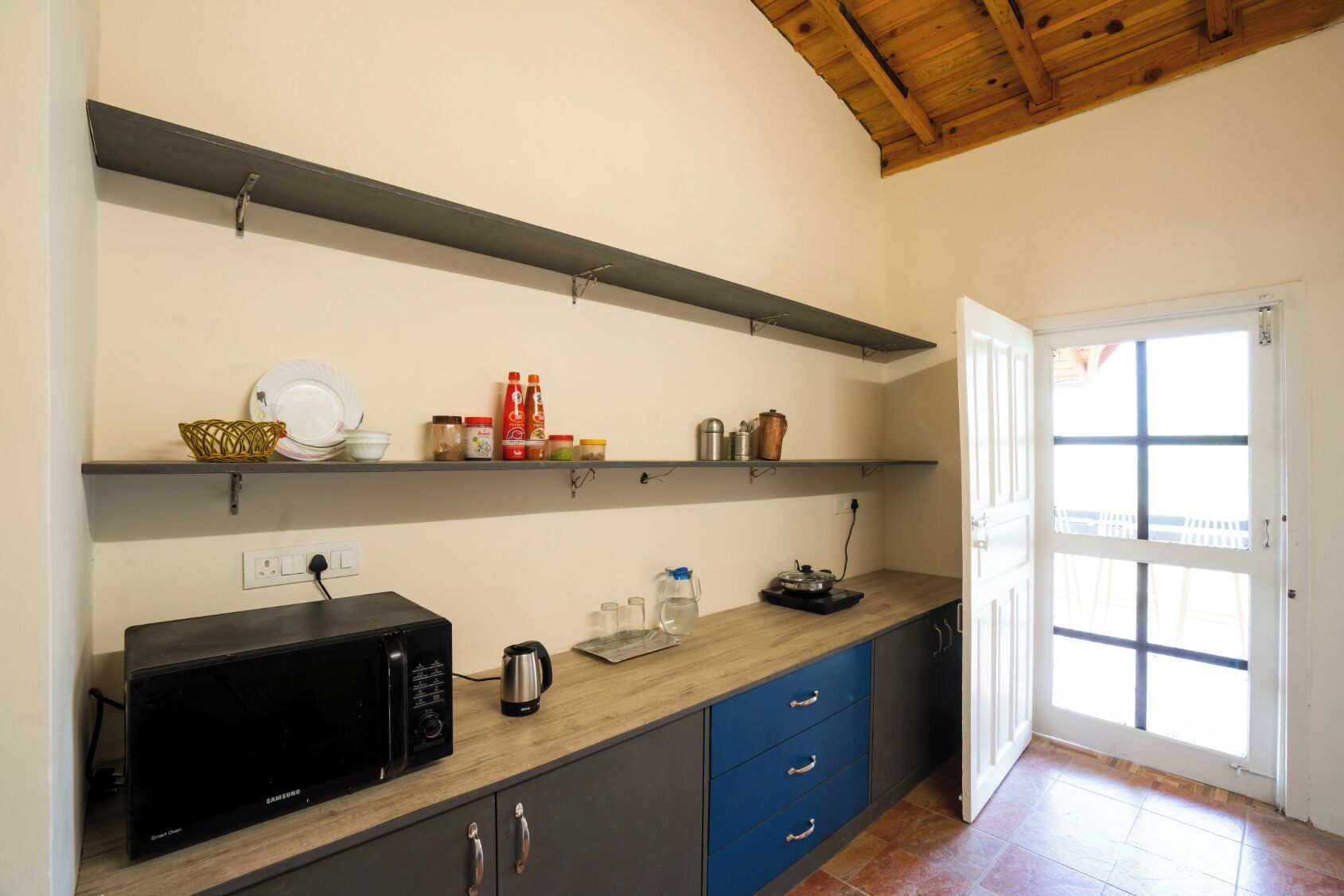A kitchen with a microwave, kettle, and various kitchen items on shelves. There is a door leading to a balcony.
