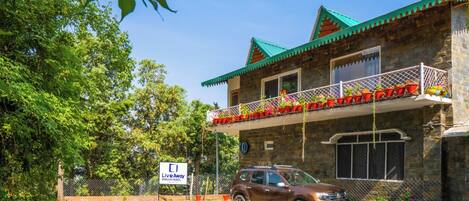A stone building with a green roof, a balcony with red flower pots, and a parked SUV.