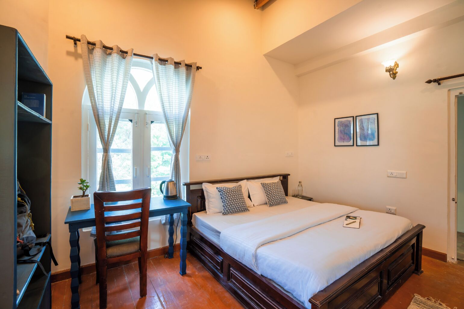 Una camera da letto con un letto di legno, una scrivania e una sedia. Si vede una finestra con tende, una lampada a parete e due quadri incorniciati.