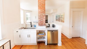 A modern kitchen with white cabinetry, a brick column, stainless steel appliances, and wooden flooring.