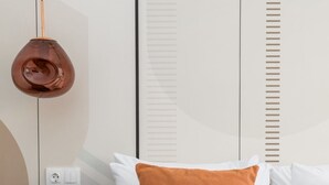 A neatly made bed with white and orange pillows, a round table with a pair of glasses, and a pendant light hanging from the ceiling. - Aurora by halu (Thessaloniki)