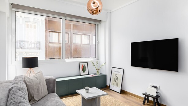 A modern living room with a grey sofa, a small white coffee table, a large window with a view of buildings, a flat-screen TV mounted on the wall, and a black lamp on a stand. - Aurora by halu (Thessaloniki)