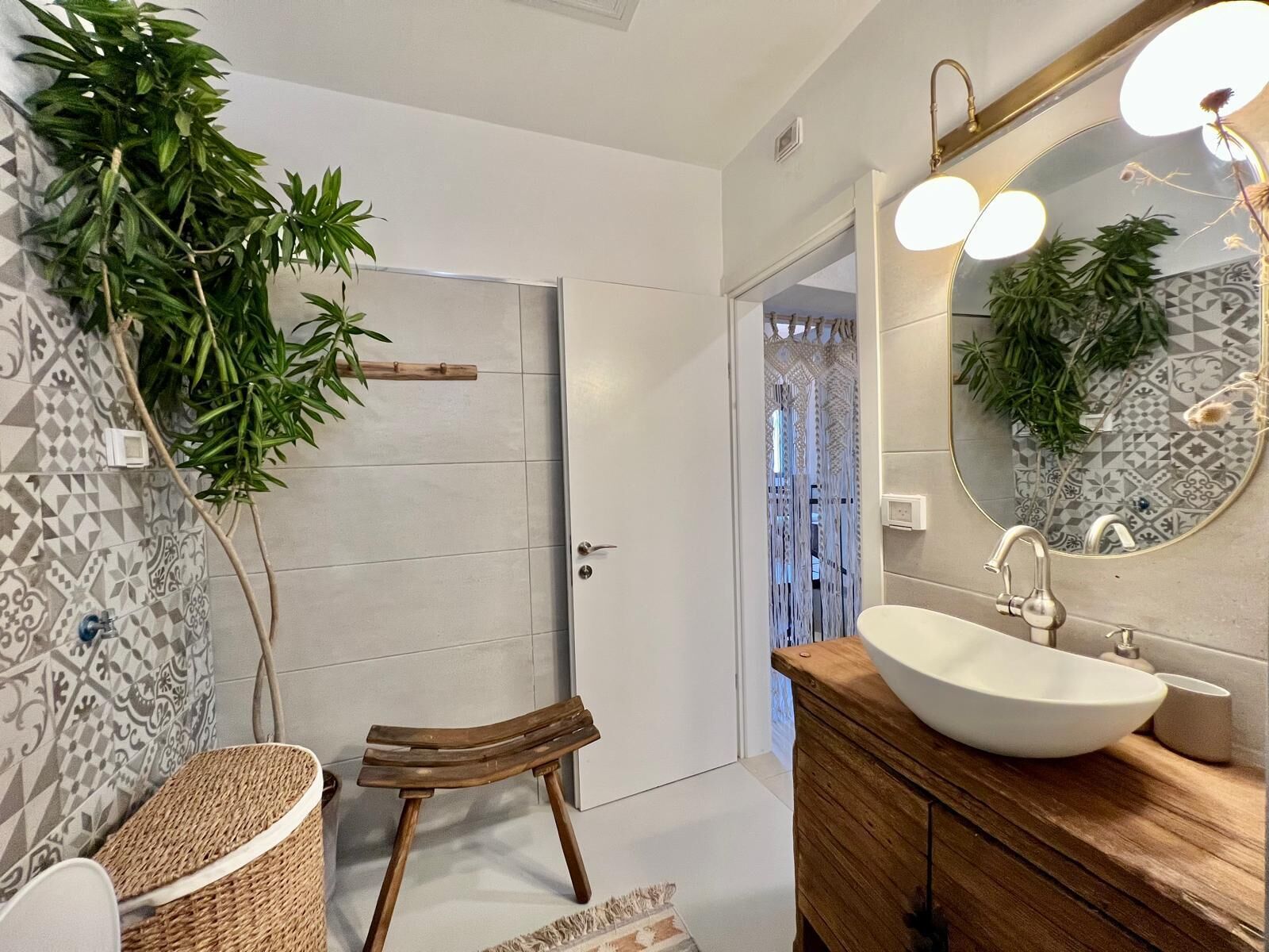 Une salle de bain avec un grand miroir, un lavabo blanc et un meuble-lavabo en bois. On y trouve une porte ornée d’un motif géométrique sur le mur, et une plante sur le côté gauche.