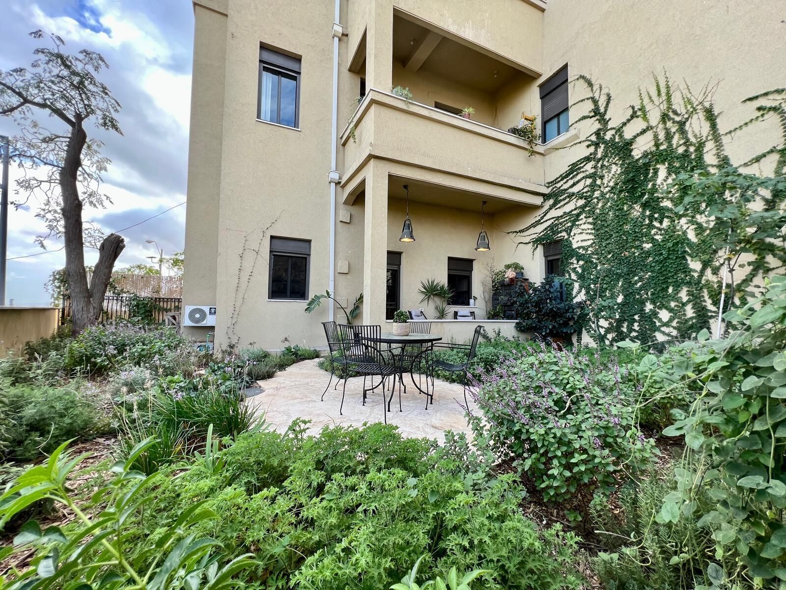 A building with a patio area featuring a table and chairs, surrounded by lush greenery and a tree.