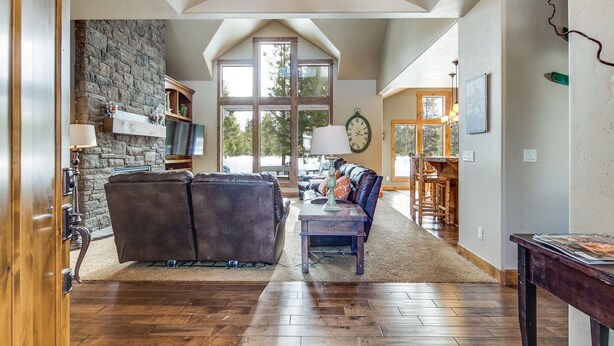 A spacious living room with a stone fireplace, leather sofas, a wooden dining table, and large windows offering a view of the outdoors. - 56540 Sunstone Loop- Caldera Springs - A/C, Hot Tub, Gas Fire Pit! (Sunriver)