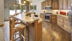 A kitchen with wooden cabinets, a granite countertop, a built-in island with bar stools, a stainless steel refrigerator, and a large window overlooking a snowy landscape. - 56540 Sunstone Loop- Caldera Springs - A/C, Hot Tub, Gas Fire Pit! (Sunriver)