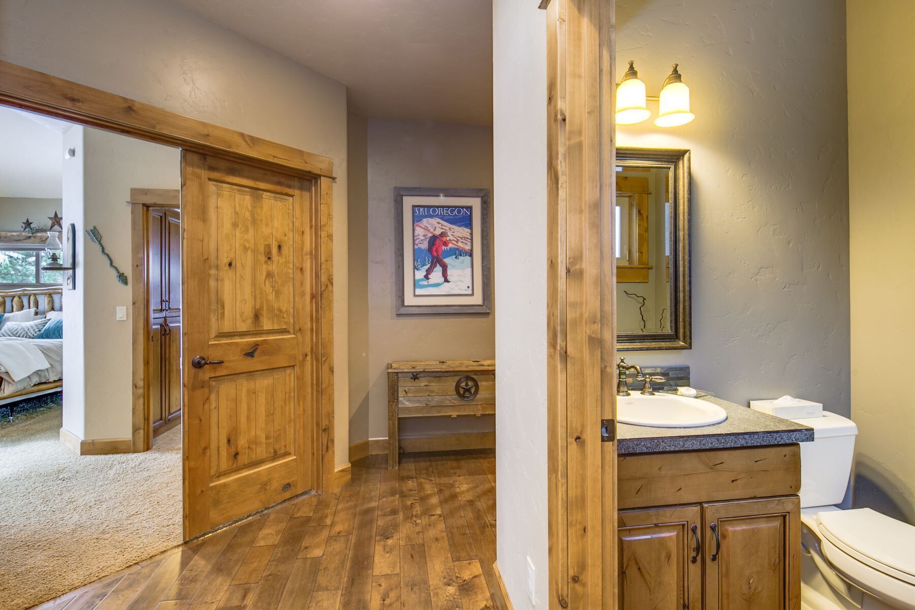 Une porte en bois donne sur une salle de bain équipée d’un lavabo, d’un miroir et d’un tableau encadré accroché au mur.