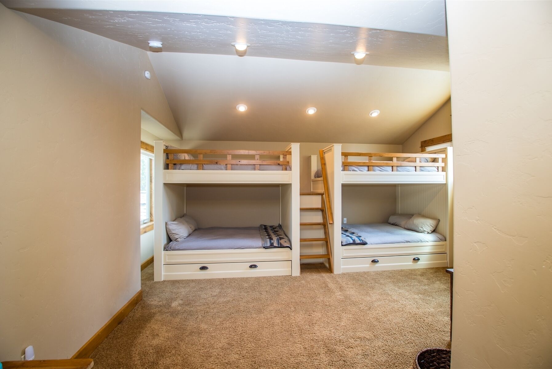 A room with two bunk beds, each with a small drawer below. The room has a sloped ceiling with recessed lighting.