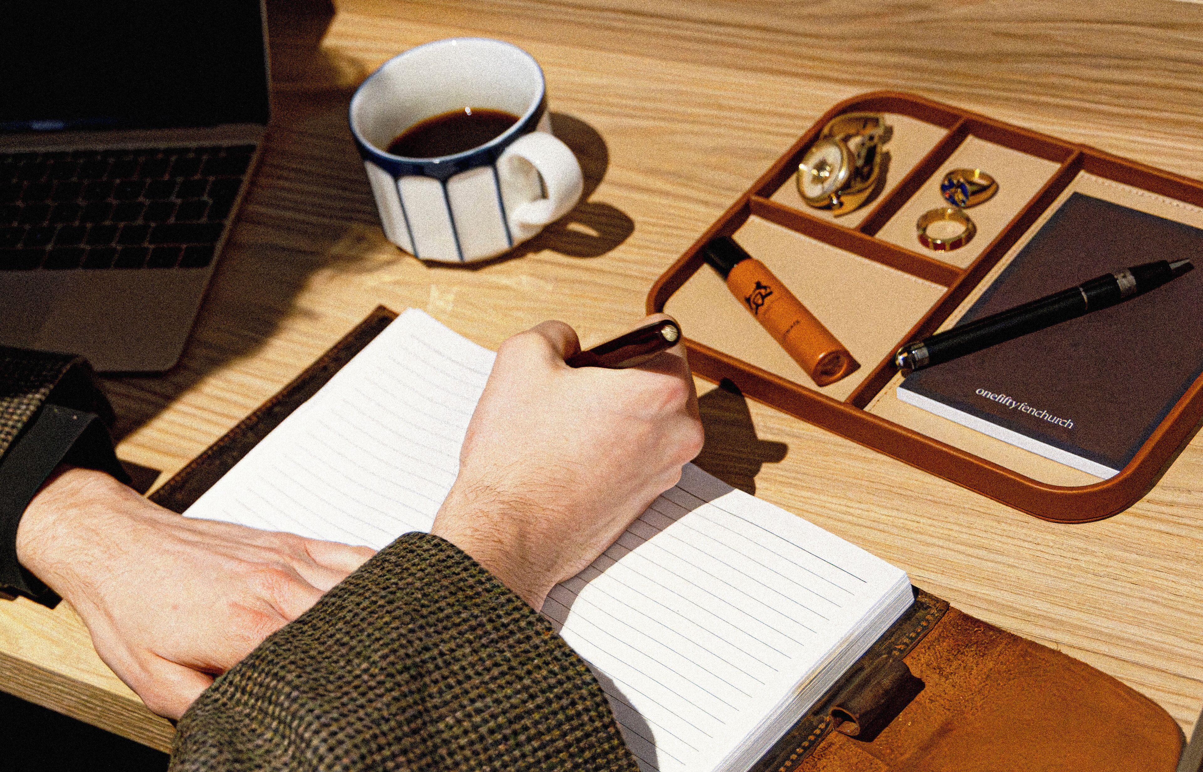 Una persona escribiendo en un cuaderno con bolígrafo, una taza de café y un portalápices con aros sobre una mesa de madera.