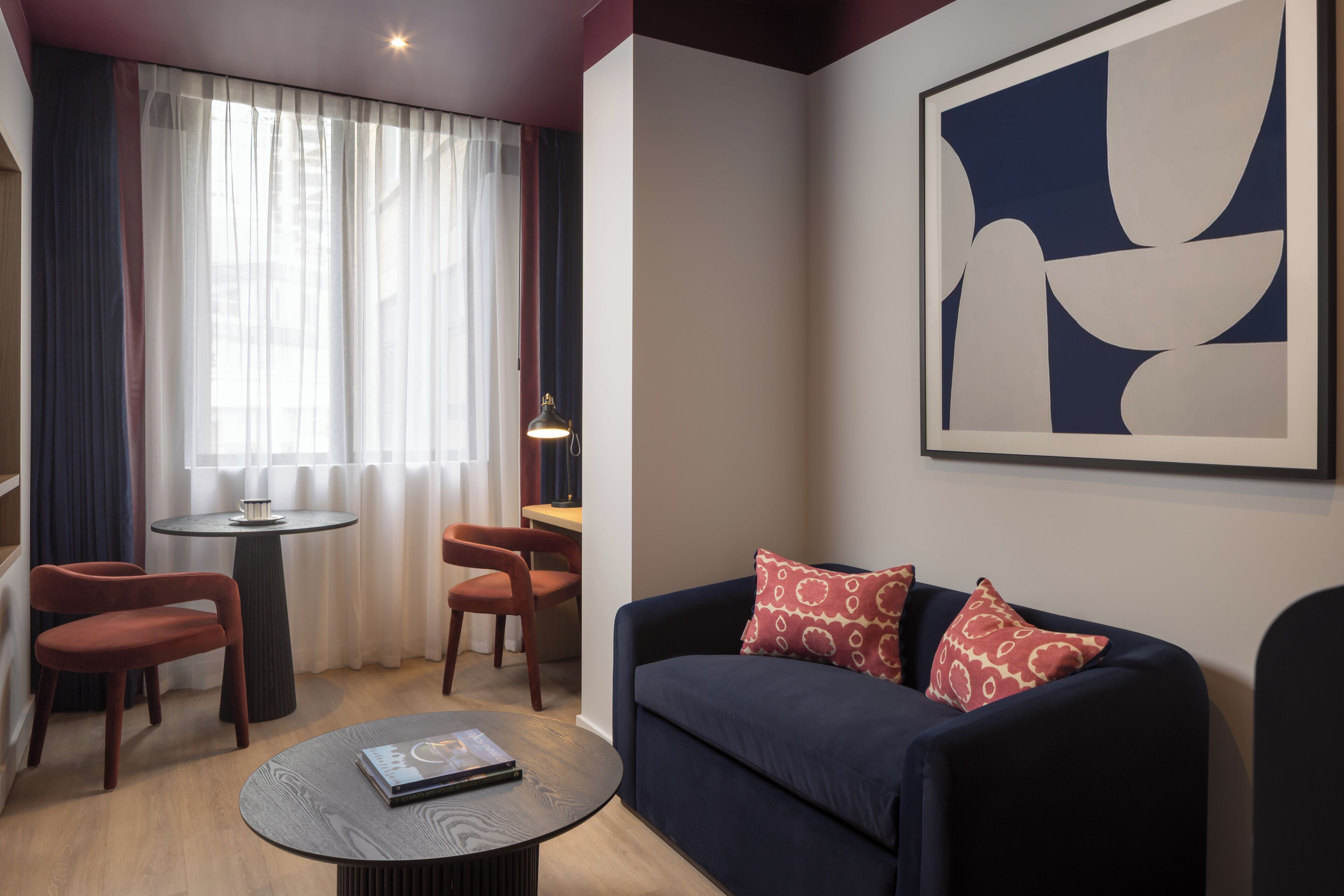 Una sala moderna con un sofá azul oscuro, almohadones estampados en rojo, dos sillas de madera, una mesita redonda con un libro y un cuadro abstracto grande en la pared.