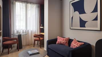 Una sala moderna con un sofá azul oscuro, almohadones estampados en rojo, dos sillas de madera, una mesita redonda con un libro y un cuadro abstracto grande en la pared.