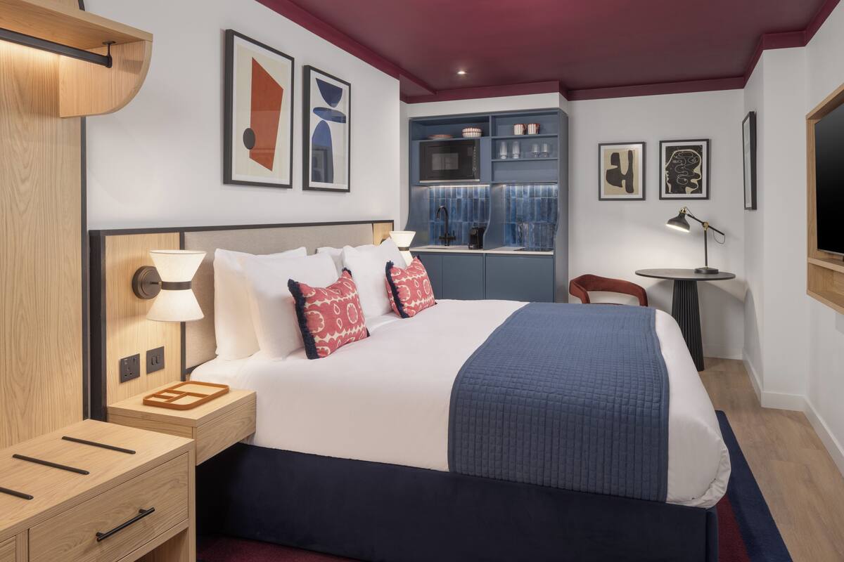 A hotel room with a bed, bedside tables, a desk, a chair, a small table, and artwork on the walls.