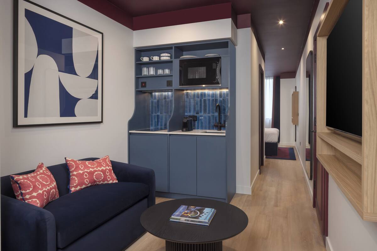 A modern living room with a dark blue sofa, a round coffee table, and a kitchen area with blue cabinets and a microwave.