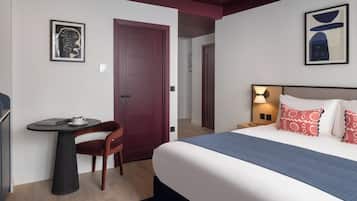 A modern hotel room with a large bed, a small round table with a chair, a door, and two framed artworks on the walls.