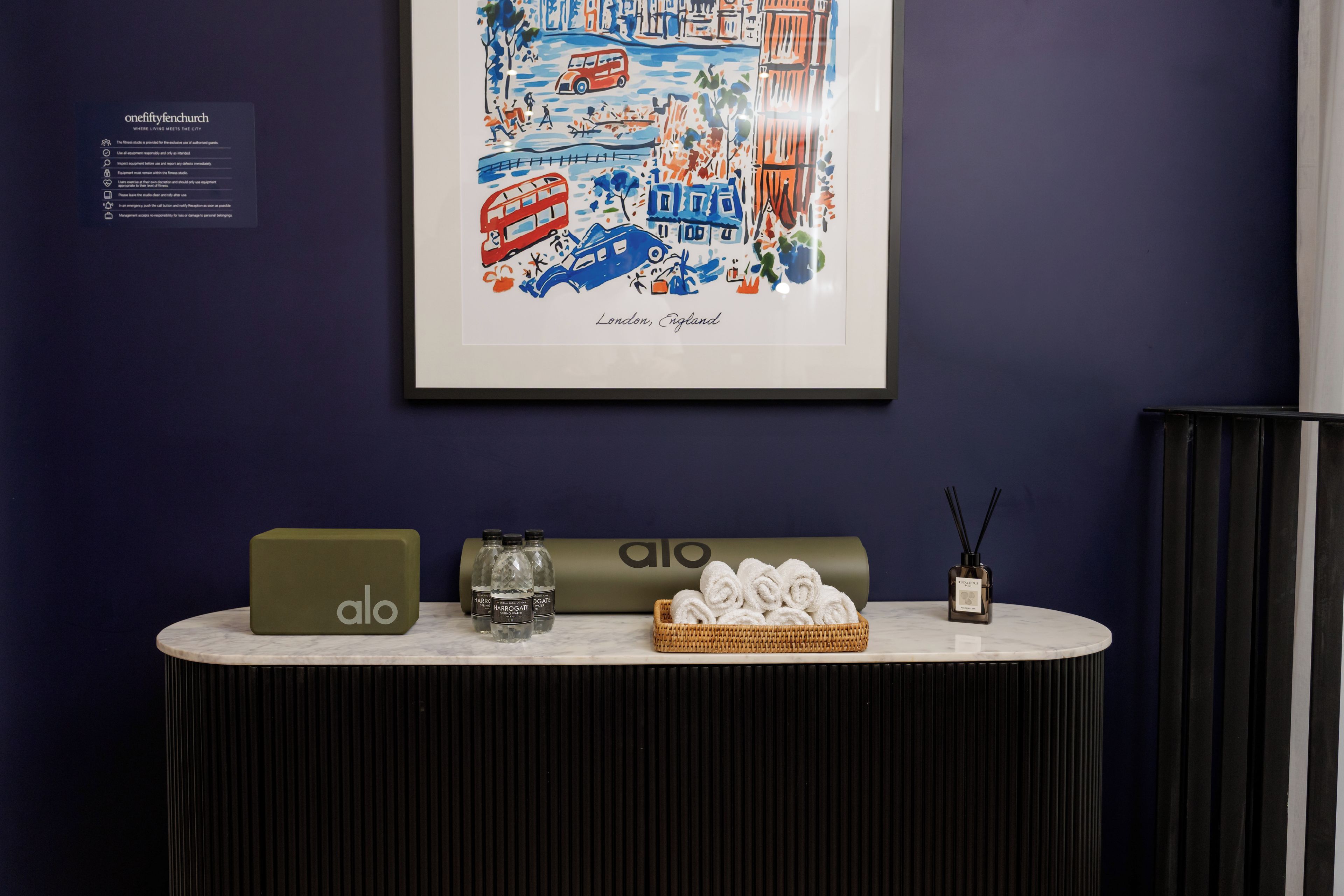 Obra enquadrada de Londres, Inglaterra, em uma parede escura. Abaixo, uma mesa com um laptop verde, garrafas de água, um difusor e toalhas enroladas, tudo sobre um fundo escuro.