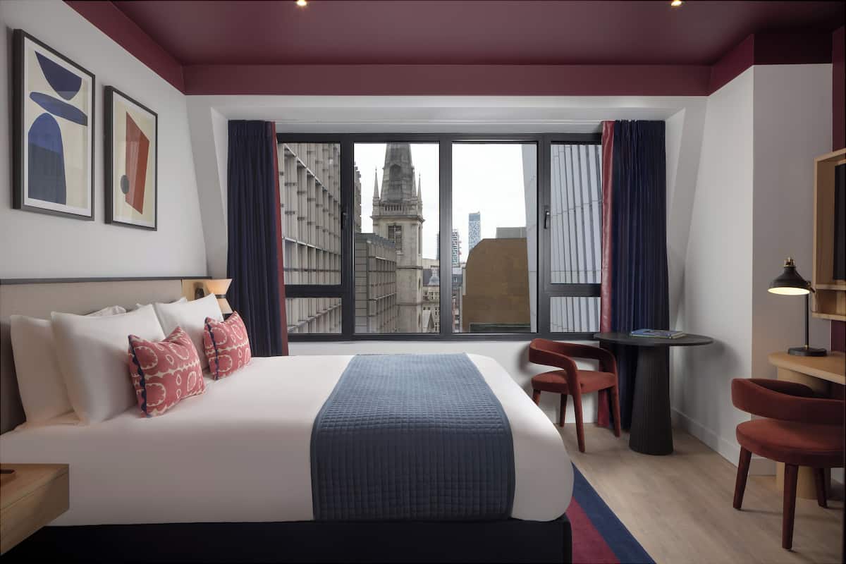 A hotel room with a large bed, a desk, two chairs, and a view of a cityscape through large windows.