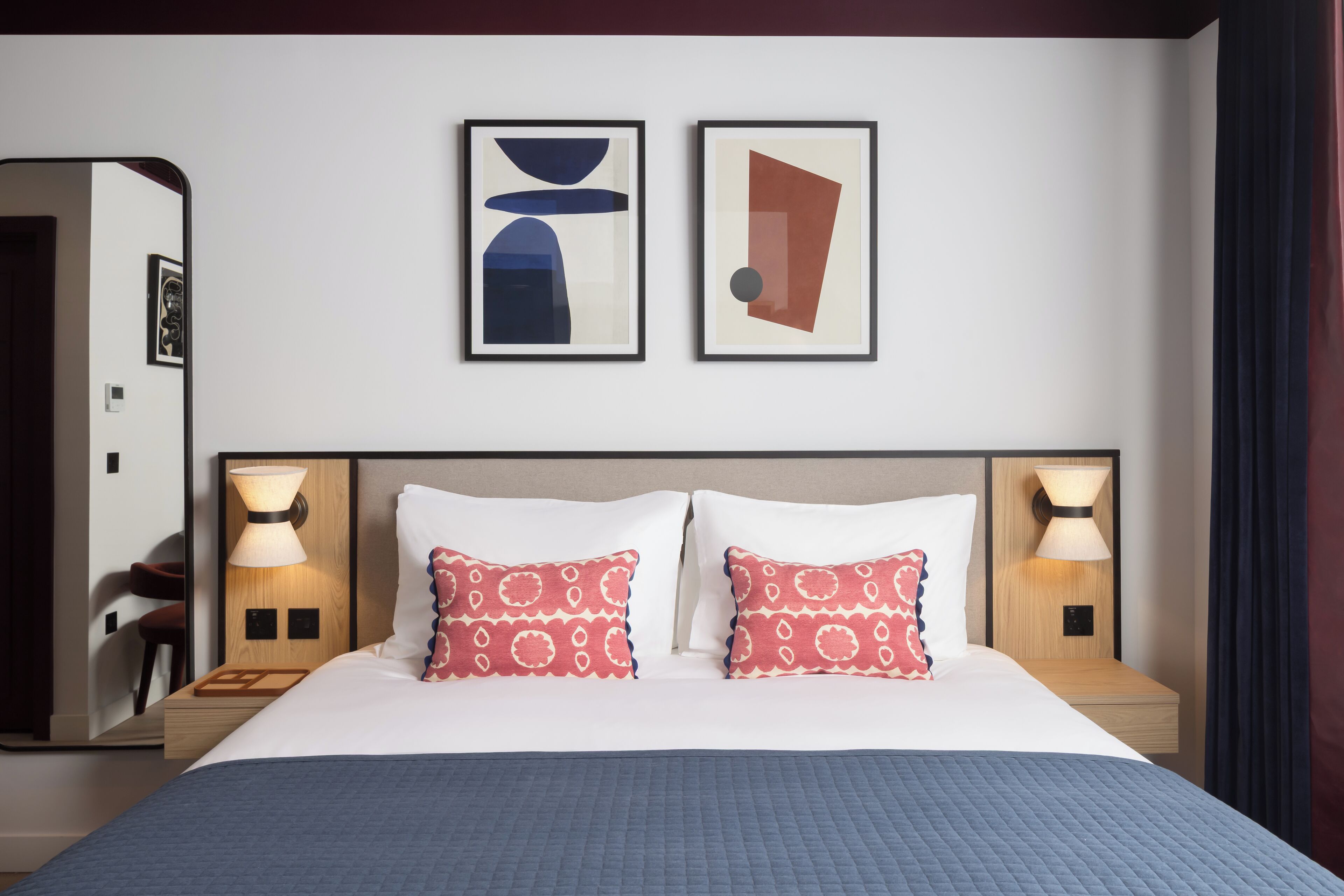 Quarto de hotel com uma cama grande, duas luminárias de cabeceira e duas obras de arte abstratas emolduradas na parede.