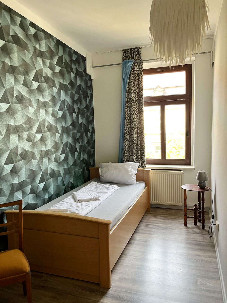 A bedroom with a geometric-patterned wall, a bed with white linens, a wooden chair, a small table with a lamp, and a window with patterned curtains.