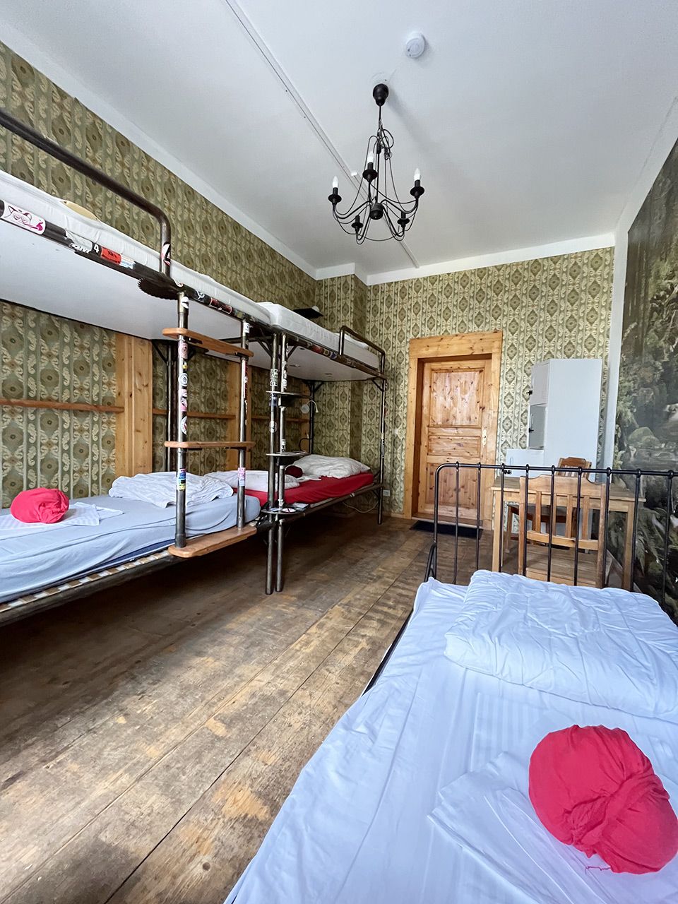 A dormitory room with bunk beds, a wooden floor, a chandelier, a whiteboard, and a door.
